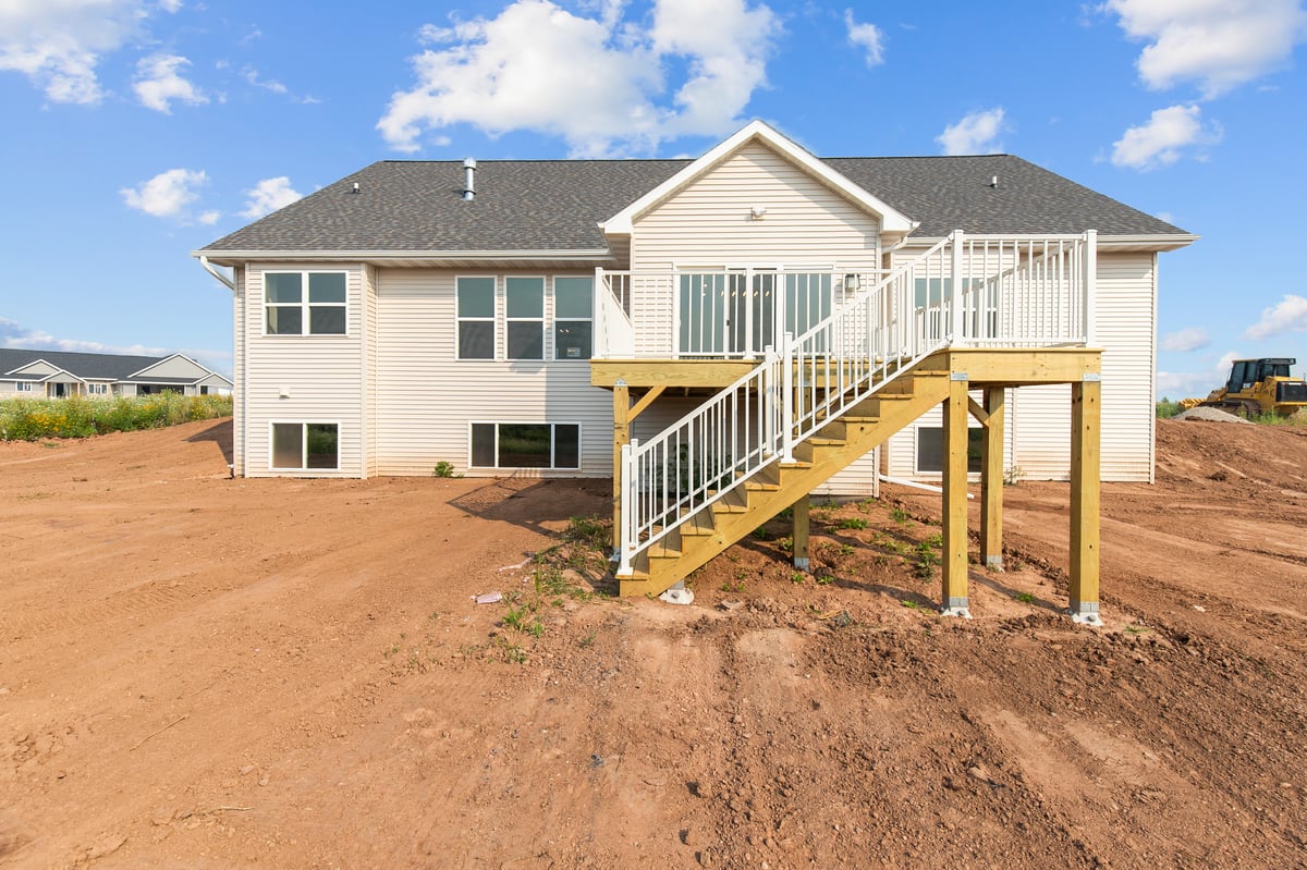 Appleton, WI rear exterior of home with elevated wood deck, white railing, staircase access, and unfinished yard under blue sky