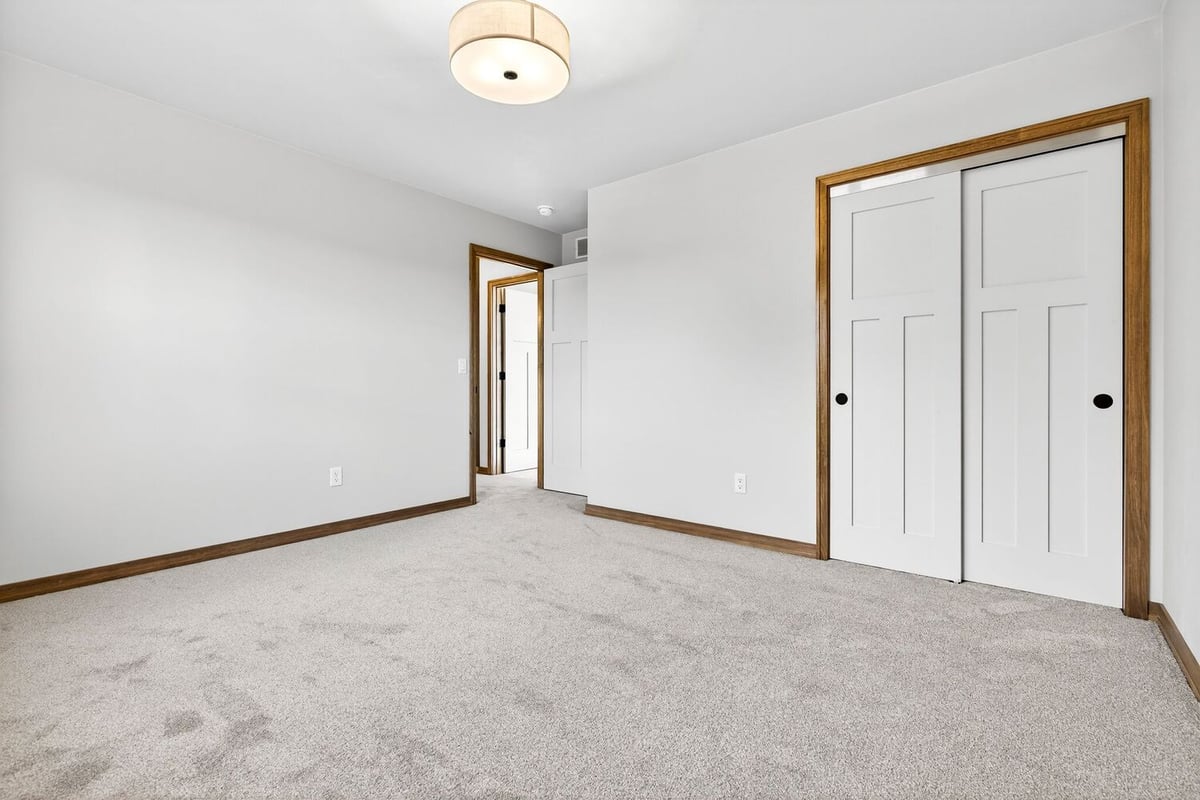 Empty carpeted bedroom with neutral walls, wood trim, ceiling light, and double closet doors in a Fox Cities home