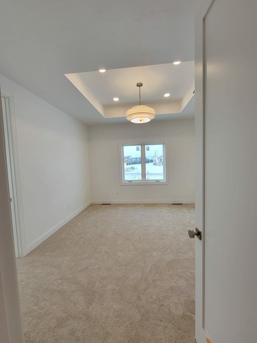 Bright carpeted bedroom with tray ceiling, recessed lighting, and modern pendant light, located in the Fox Cities