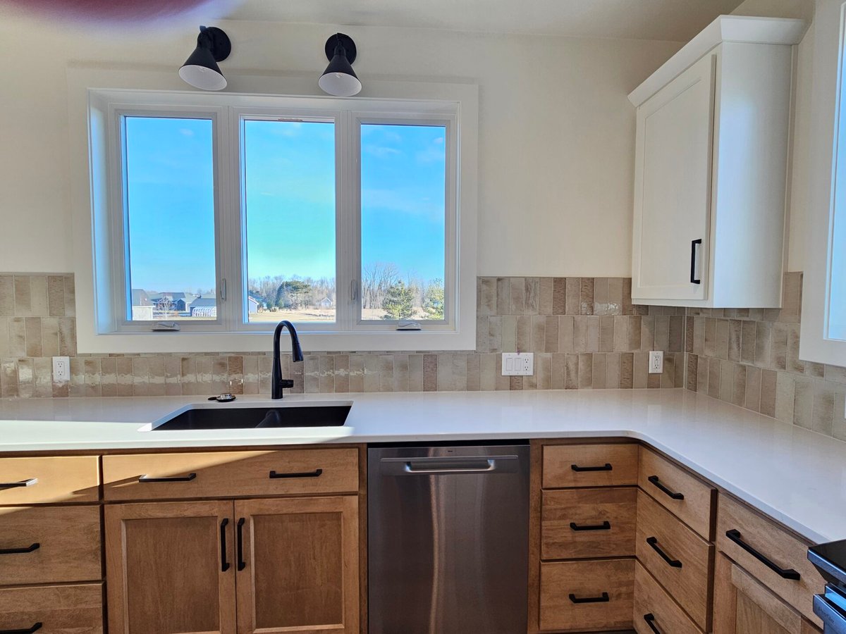 Kitchen sink area with wood cabinets, white countertops, black fixtures, and large window, located in the Fox Cities