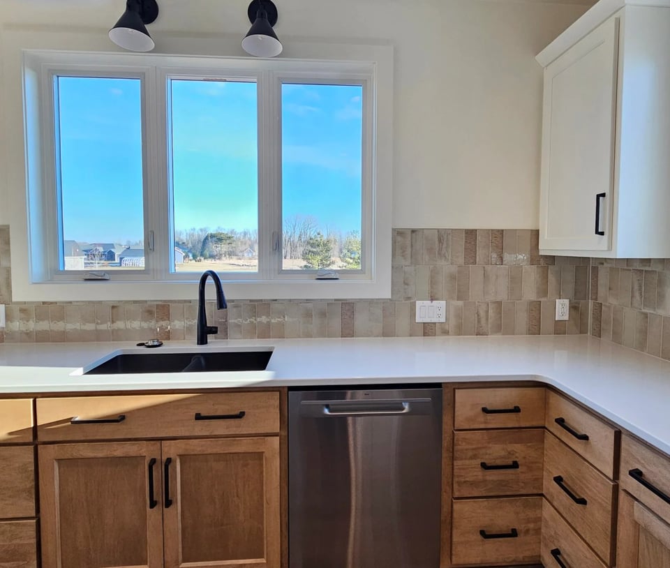 Kitchen sink area with wood cabinets, white countertops, black fixtures, and large window, located in the Fox Cities.